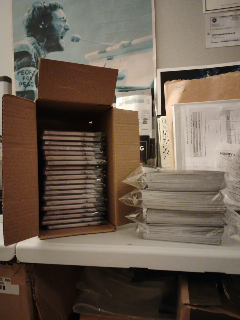 A box packed with CDs in jewel cases next to stacks of shrink-wrapped discs